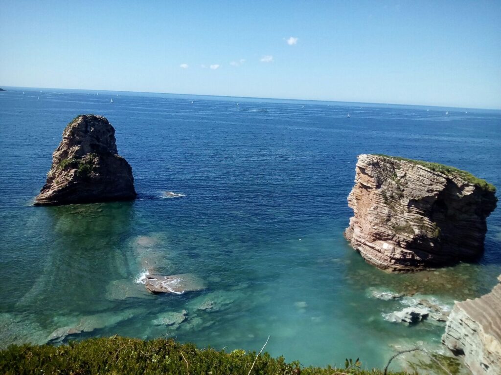 Two large sea rocks rise from turquoise water with a clear blue sky and distant sailboats along the horizon.