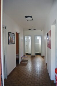 White hallway with brown tile floor, framed artwork on the left, several doors along the corridor, and a red fire extinguisher mounted on the right wall near the end of the hall.