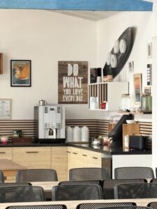 Cafe counter with an espresso machine, cups, and shelves, plus wall art in the background and metal chairs in the foreground.