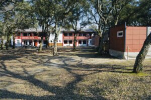 Two-story white building with red trim tucked among large trees in a courtyard, with benches and table seating outside and a red-orange modular structure to the right.