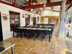 Outdoor beach club dining area with long wooden tables and black plastic chairs under a covered patio with red beams.