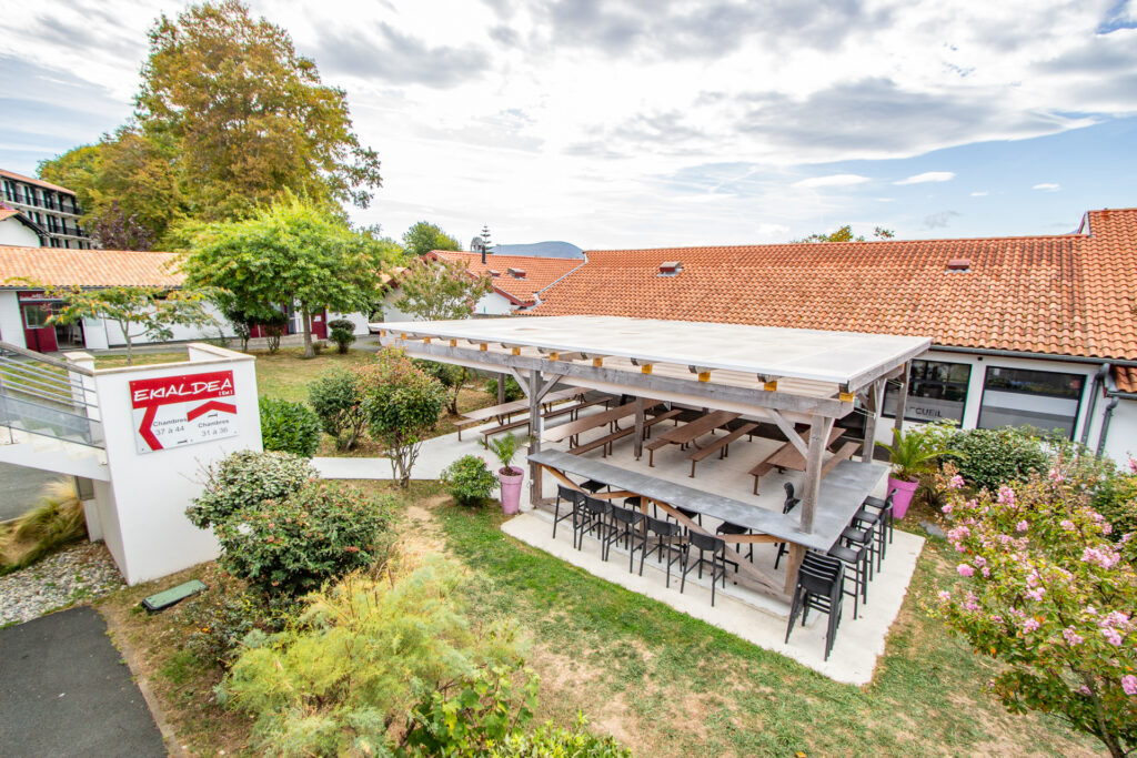 Outdoor courtyard with a large wooden pergola and long picnic-style benches beside red-tiled roofs and flowering plants